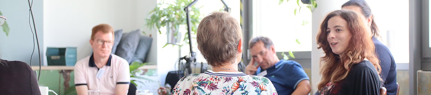 Podcast Workshop Mehrere Menschen sitzen in einem hellen Raum um einen Tisch herum und tauschen sich aus.